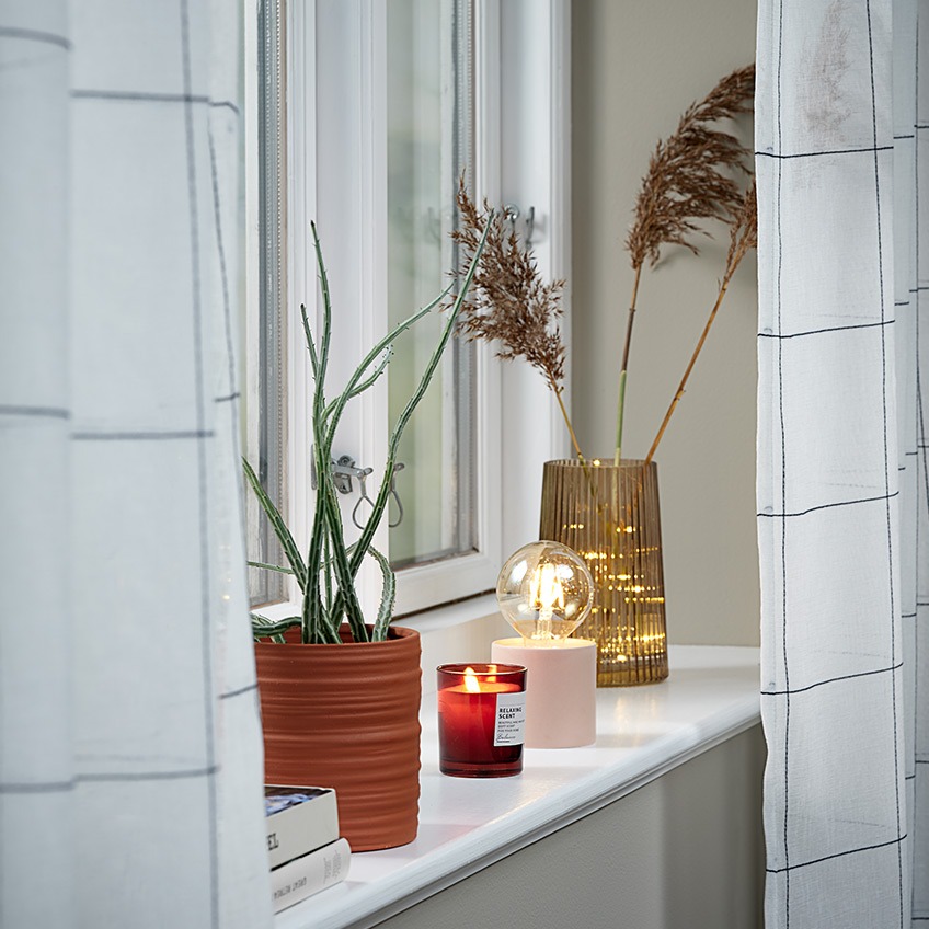 Lightweight curtains in a window with plant pot, scented candle, battery lamp and vase in the windowsill