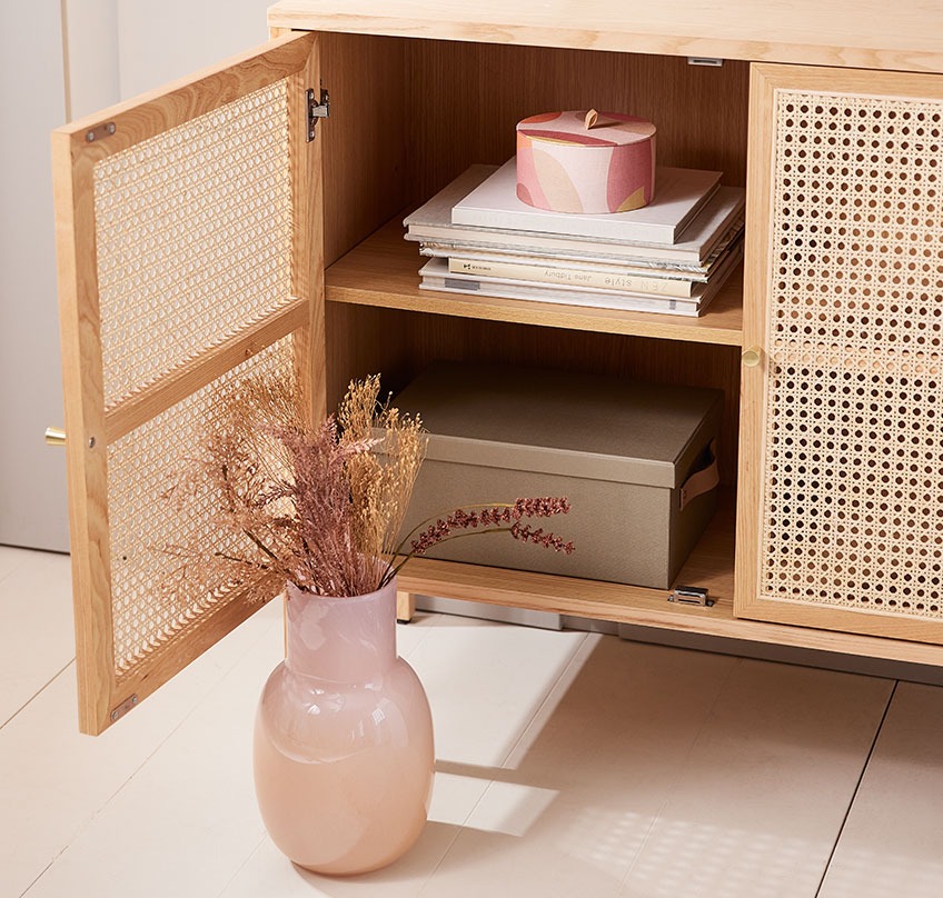 Mouth blown vase with a bouquet of artificial flowers in front of sideboard with an open door and a round storage box inside 